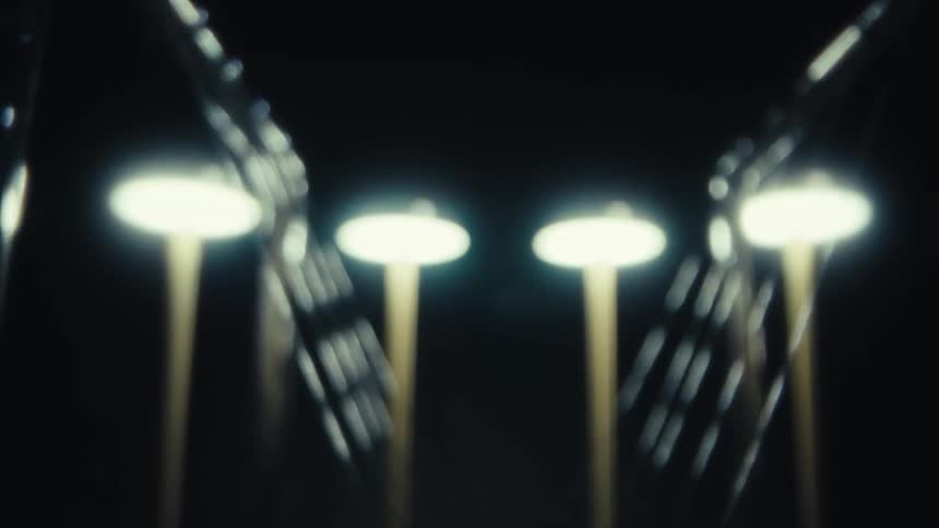 Pendulum balls of a newtons cradle hang beneath bright overhead lights their blurred motion and glowing highlights creating an abstract rhythmic pattern against a dark background