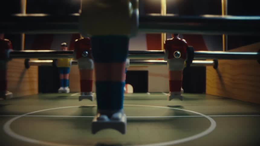 A foosball table midgame with painted player figures in colorful uniforms poised around a small blackandwhite ball under warm moody overhead lights creating a dramatic almost arenalike atmosphere despite the miniature scale