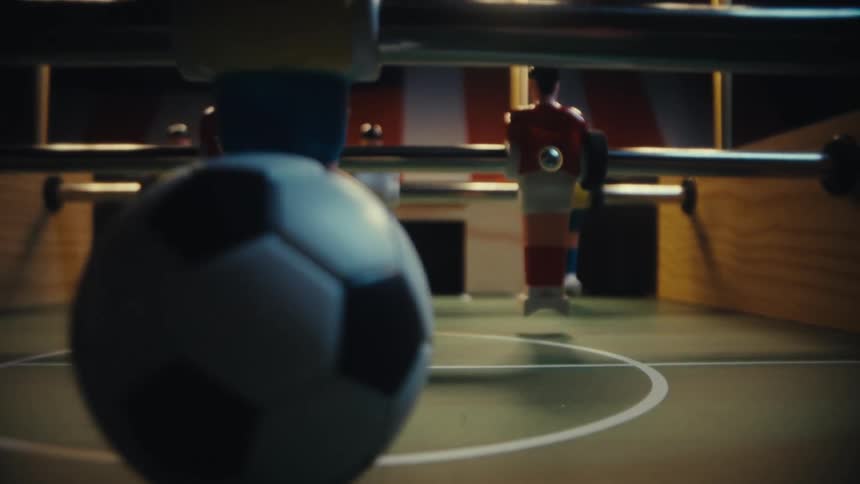 A lowangle closeup shot captures a fastpaced game of foosball with the camera tracking a small soccer ball as it zips across the green felt pitch between the rigid painted plastic players under warm overhead lighting