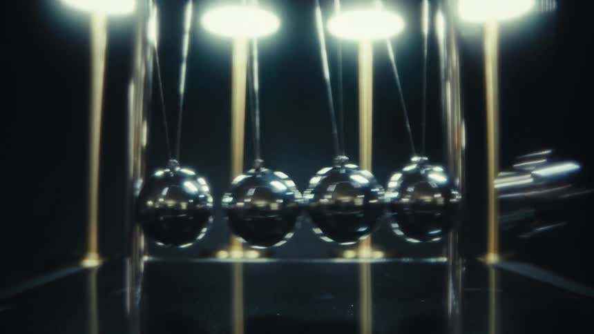 A newtons cradle sits in a dark spotlighted environment showing four shiny metal spheres coming to life as the outer balls begin to swing and strike the center pair demonstrating the continuous transfer of kinetic energy with rhythmic clicking precis