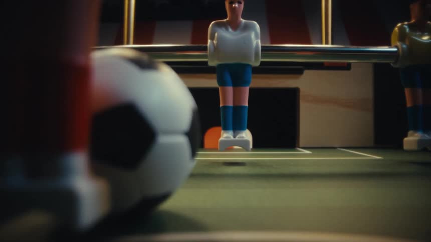 A lowangle closeup shot captures a fastpaced game of foosball with the camera tracking a small soccer ball as it zips across the green felt pitch between the rigid painted plastic players under warm overhead lighting