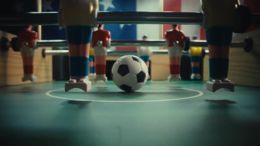 A foosball sits at the center of a green pitch framed by the plastic legs of the players and an american flag hanging in the background the camera zooms in from a lowangle perspective focusing closely on the stationary soccer ball