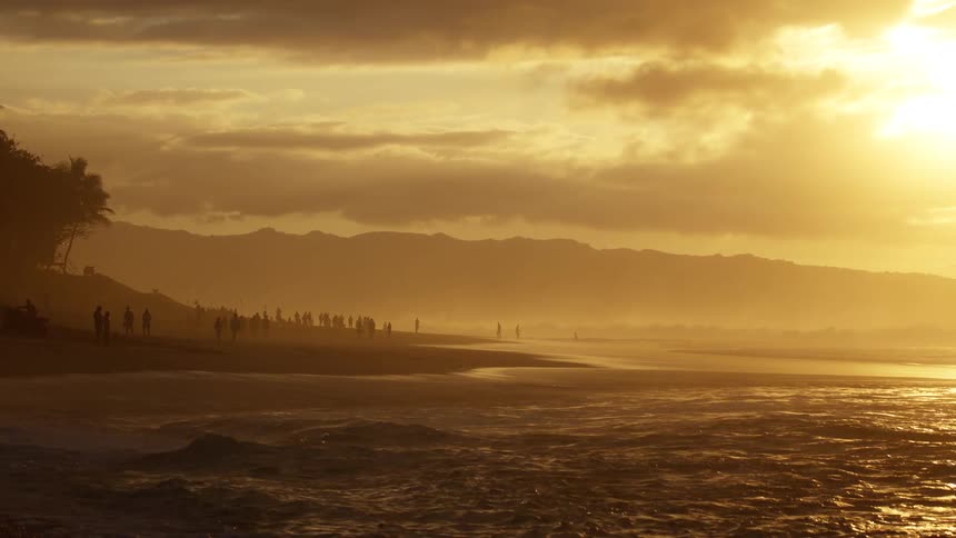 The sun sets over the ocean casting a golden glow across the water and silhouetting birds as they fly through the hazy sky the warm light reflects off the waves and onto the wet sand of a crowded beach where people are walking along the shore