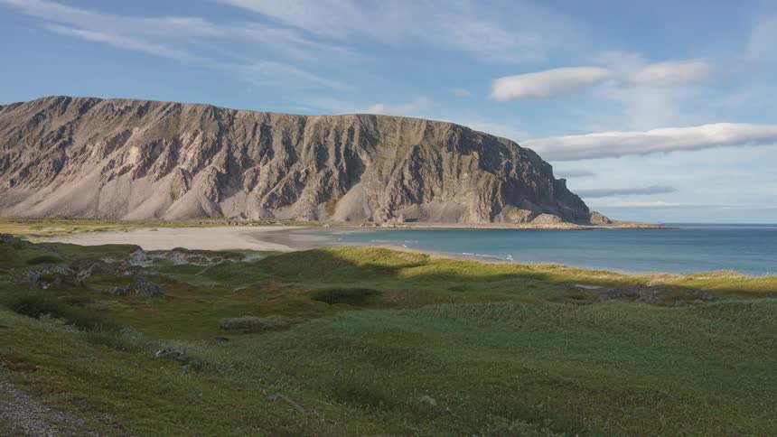 Scenic coastal landscape featuring rolling green hills a sandy beach and calm blue water at the base of a massive rocky mountain cliff under a partly cloudy sky