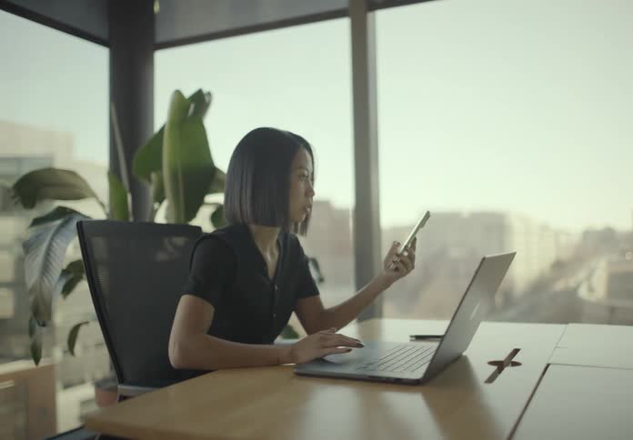 A woman sits at a wooden desk checking her smartphone while her other hand rests on a laptop displaying a colorful pie chart suggesting she is multitasking between digital analysis and mobile communication in a bright modern workspace