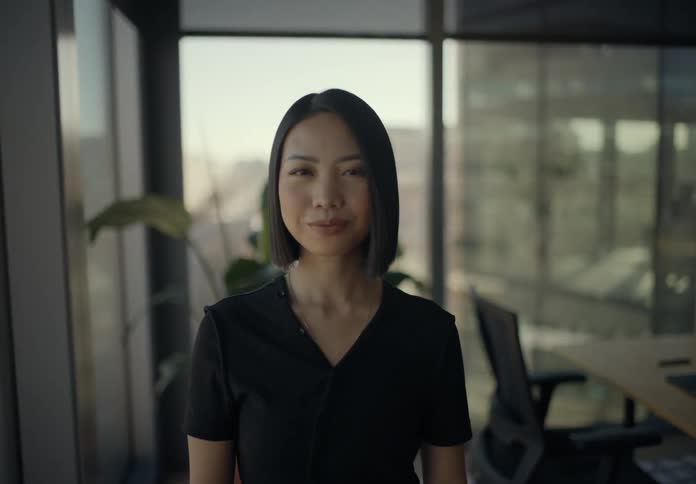 A young woman with short black hair stands in a modern glass walled office then she stops using her smartphone looks directly into the camera with a subtle smile and maintains a confident posture as the camera moves closer to her