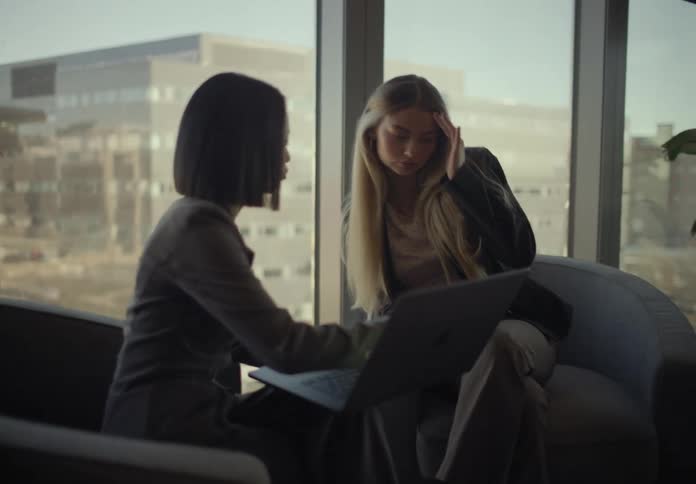 Two young adult women sitting on armchair and talking while one of them points to a laptop computer on her lap indoors during day time