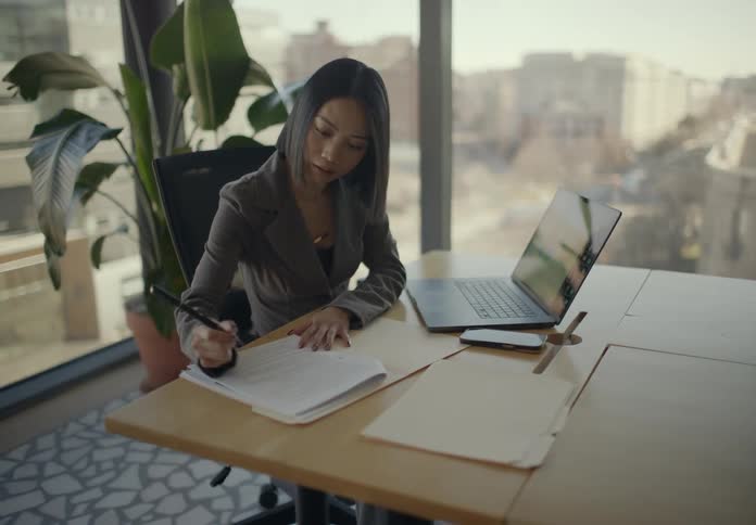 A woman in a grey blazer sits at a wooden desk in a sunlit office reviewing a colorful pie chart on her laptop before taking notes on a document and large windows behind her show a city view also a large plant stands in the corner