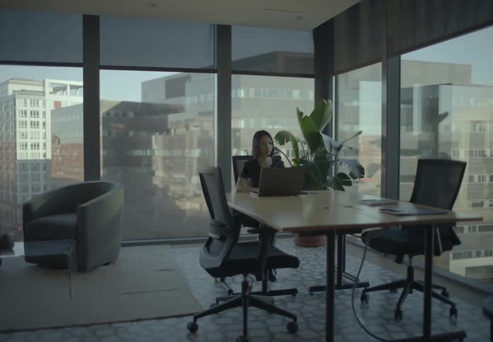 Woman working on a laptop in a corporate meeting room with table chairs and large windows in the background