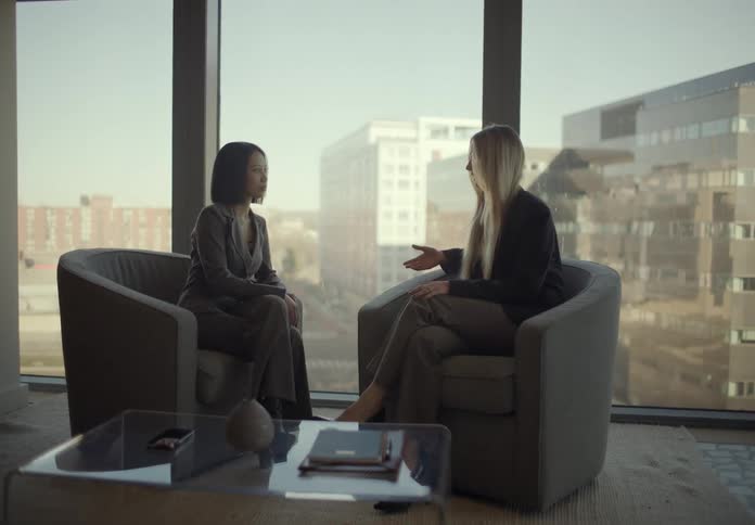 Two young adult women sitting on armchairs and talking in an office with big windows showing a metropolitan area on the background indoors during day time
