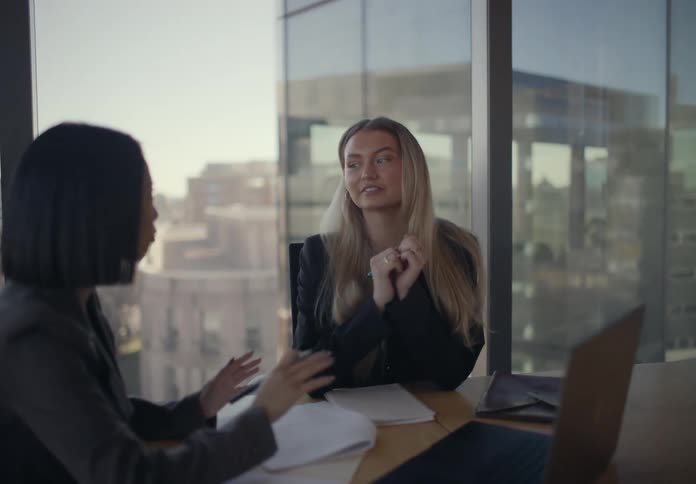 Two young adult women sitting on an office talking and pointing at the screen of a laptop computer over a window table indoors during day time