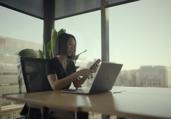 Woman sits at a table in an office working an a laptop computer and holding her phone