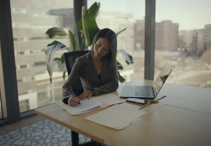 A blonde woman walks into a sunlit office to place a folder of papers on a wooden desk prompting her seated colleague to look up smile and express thanks before continuing to write