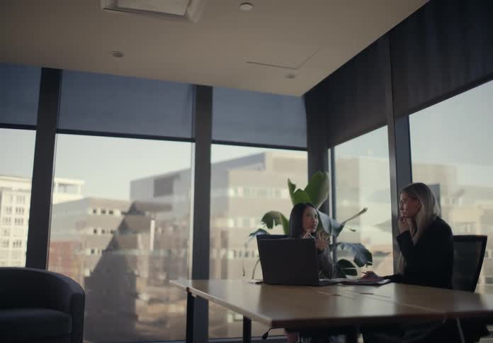 Two professional women have a serious discussion at a conference table in a modern high rise office with large windows overlooking the city