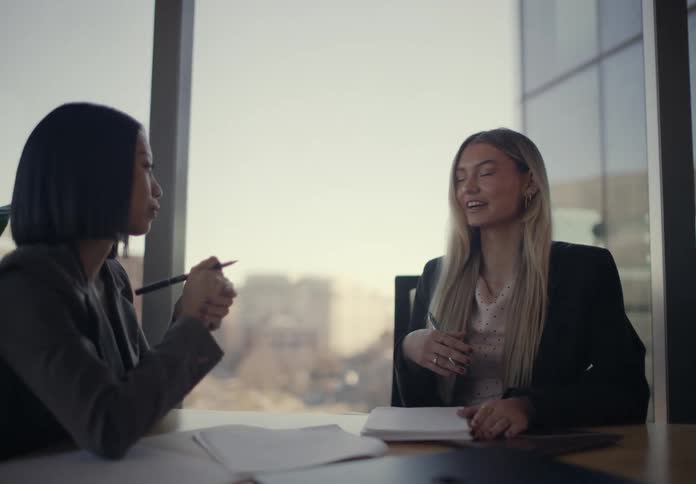 Two professional businesswomen sitting at a desk in a sunlit office engaging in a friendly and animated conversation while reviewing paperwork