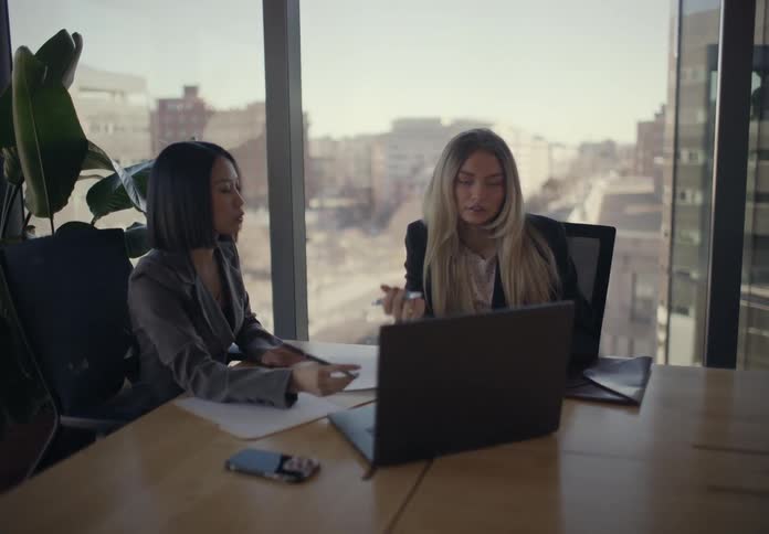 Two businesswomen collaborate at a conference table in a modern high rise office discussing work and reviewing documents on a laptop