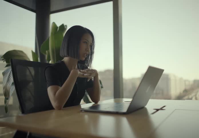Young adult woman sitting down and typing on a laptop computer over a wooden table in a room with big windows showing a metropolitan area indoors during day time