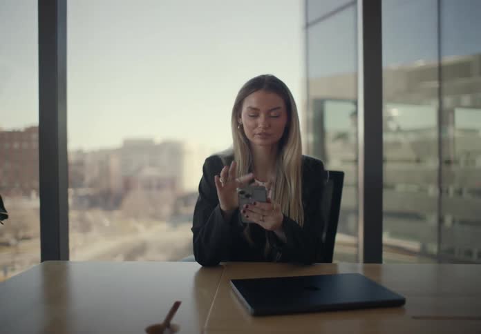 A woman in a black blazer sits at a wooden desk in a modern office closes her laptop and starts scrolling through her smartphone against a backdrop of large windows overlooking a city during the daytime