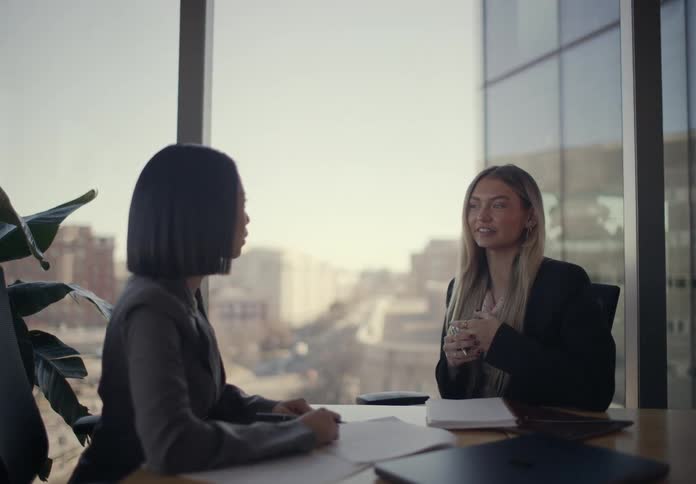 Two women sitting in an office talking over paperwork with a phone and laptop computer on the table