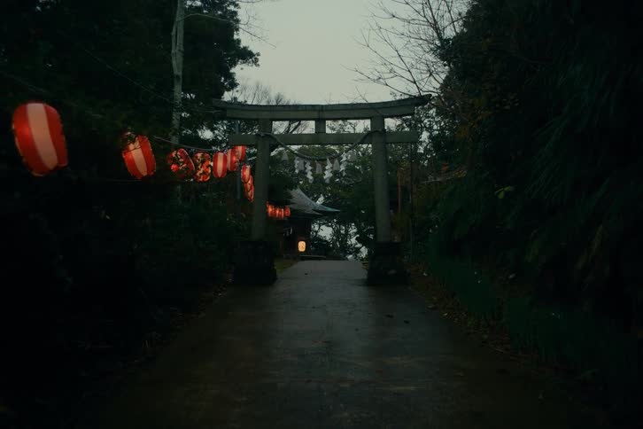 Wet pathway leading to a stone torii decorated with red lanterns in a wooded area