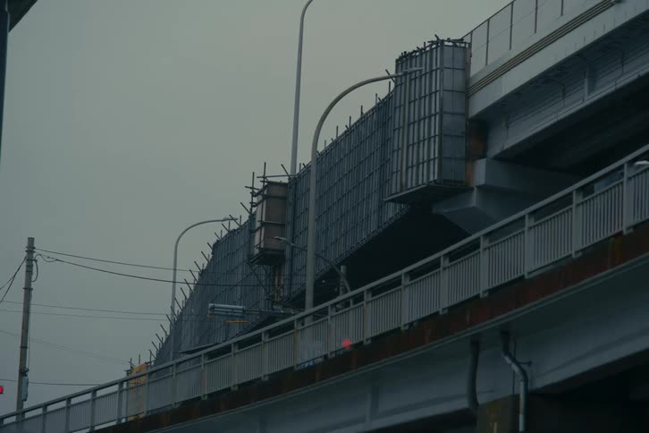 Under a gloomy overcast sky a massive concrete highway overpass undergoes construction with metal scaffolding and panels attached to its side on a lower bridge level visible through the structural supports a lone pedestrian carrying an umbrella can be