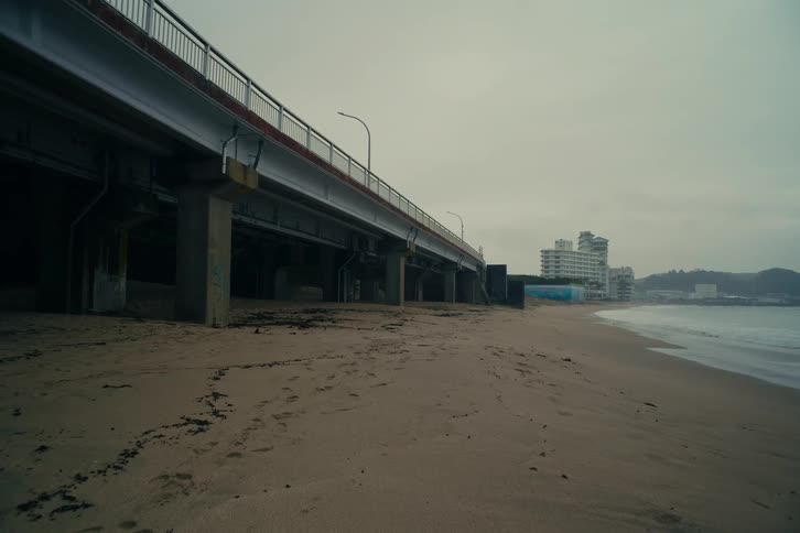 Huge concrete pillars support a highway bridge towering over a quiet sandy beach where gentle waves roll onto the shore beneath a gloomy overcast sky