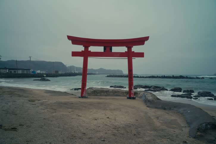 Red torii gate standing by the seaside with breaking waves and an overcast sky in the background