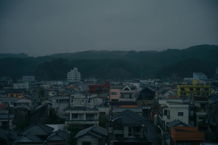 A wide highangle view captures a densely packed japanese town under a heavy overcast sky where a sea of varied gray and colorful rooftops stretches toward dark forested rolling hills in the distance