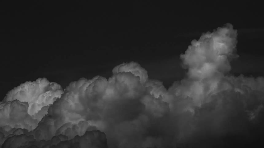 Large monochrome cumulus clouds billow and expand against a dark night sky showing constant internal movement and shifting textures as they surge upward in a dense formation