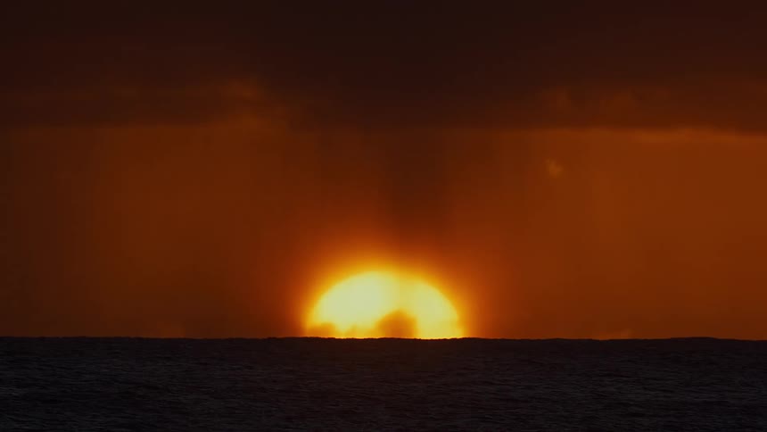 A massive glowing sun hovers on the horizon of a dark restless ocean veiled by heavy orange clouds and sweeping curtains of rain
