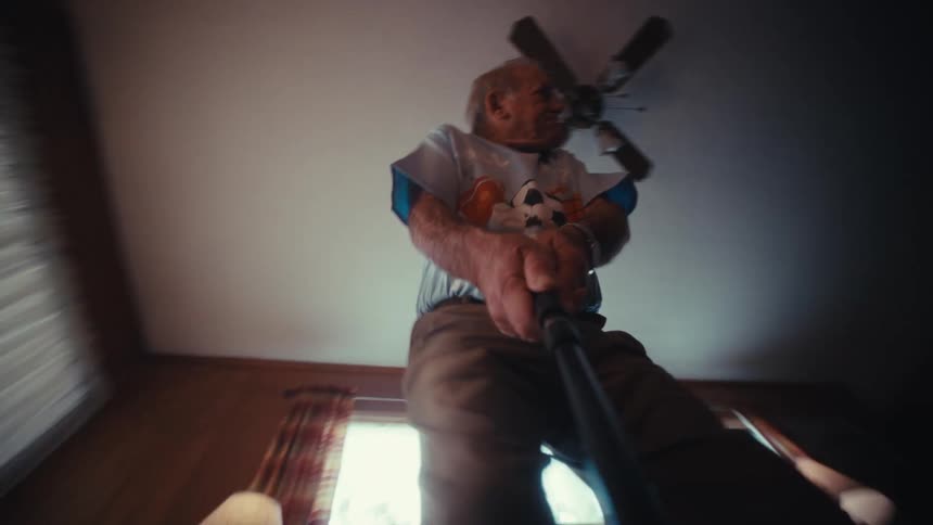 An elderly man in a soccer tshirt and khaki pants with a stick in a living room filmed from a low angle on the floor looking up as he moves back and forth under a ceiling fan near curtained windows