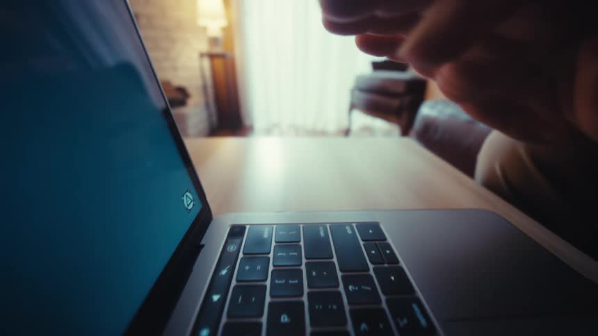 A closeup shot shows a mans hand typing on a laptop keyboard and using the trackpad while sitting in a dimly lit living room with a leather chair and a window in the background