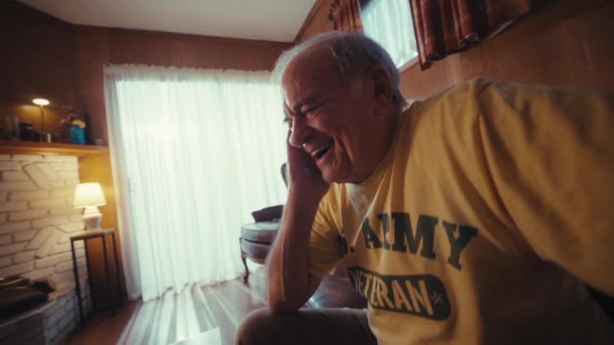 An elderly man sitting in his living room overly excited and laughing as he looks at a laptop on the coffee table in front of him