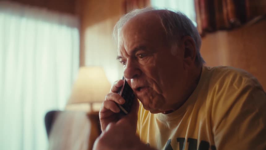 Seated in a cozy woodpaneled living room an elderly man in a yellow shirt gestures toward his open laptop while engaged in an animated conversation on his mobile phone