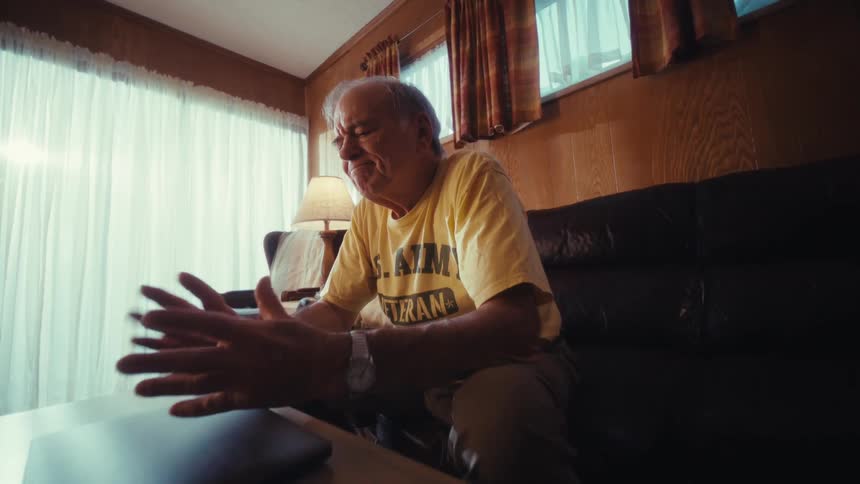 An elderly veteran in a yellow shirt sits on a leather sofa finishes using his laptop and closes the lid before staring forward with a somber expression in a living room