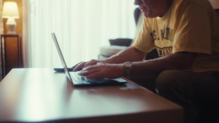 Senior man sitting on a leather sofa and typing on a laptop computer in a home living room with a closeup on his hands