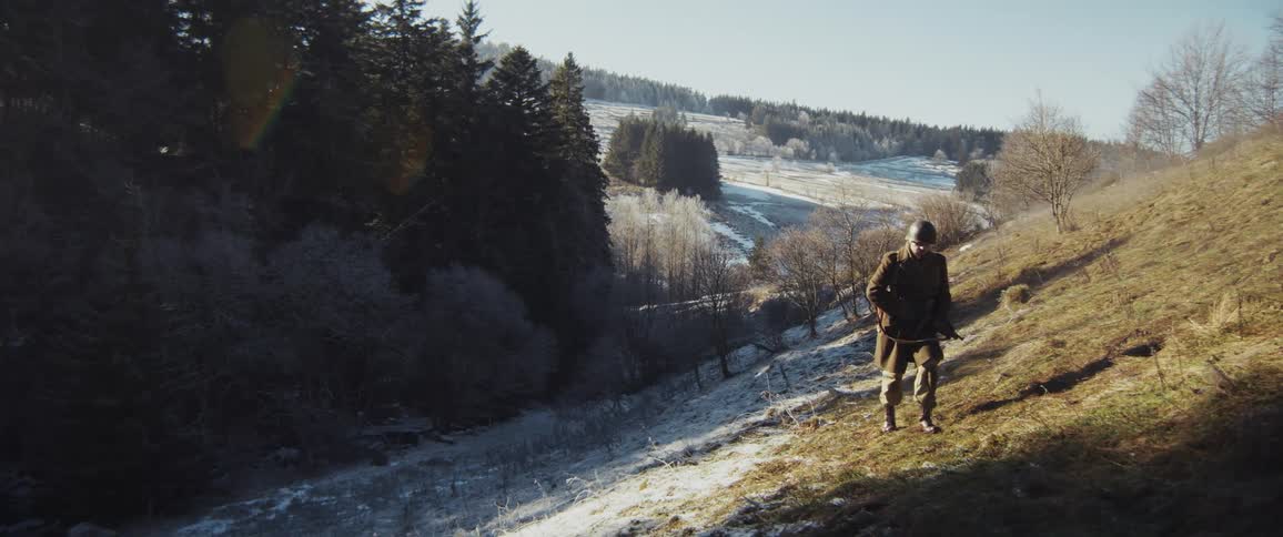 A soldier in a heavy winter coat and helmet walks down a frost covered hillside toward a valley