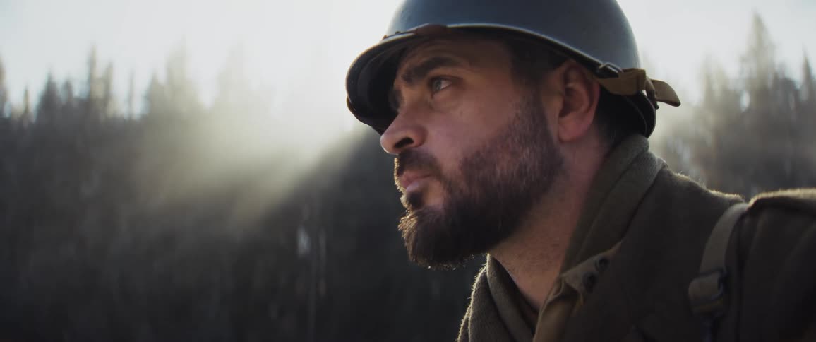 Bearded soldier wearing a steel helmet and wool uniform stands in a misty forest looking around with a heavy expression as sunlight filters through the trees behind him