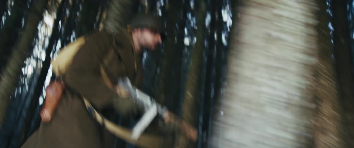 A soldier in a wool overcoat and helmet runs through a dense forest while holding a rifle