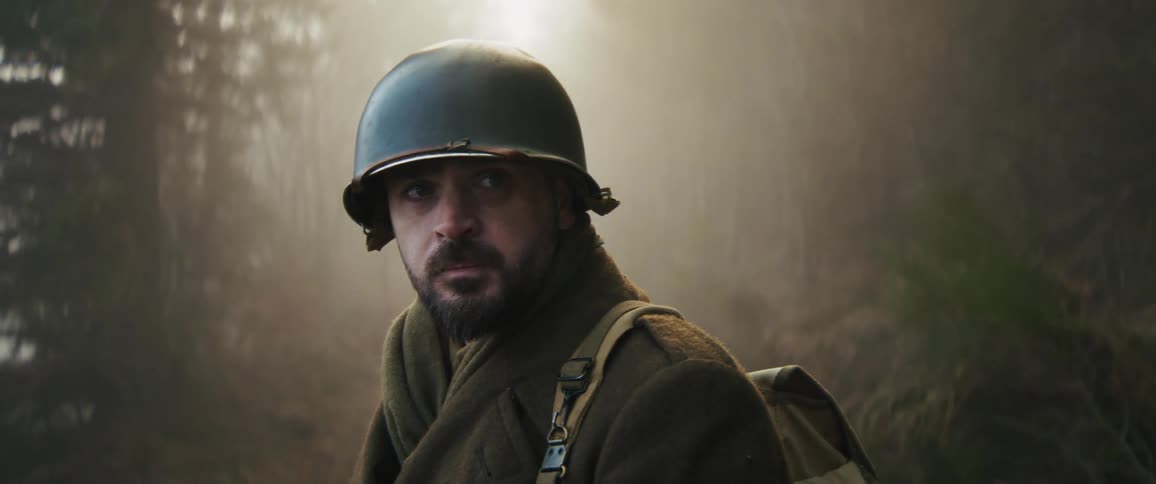 A male soldier with a beard walks through a foggy forest then stops and turns his head to look behind him with a concerned expression