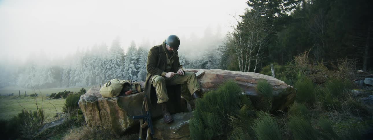 A male soldier sits on a large rock in a foggy frostcovered forest takes a cigarette from his pocket lights it and looks around at the misty trees while his rifle and bag rest beside him