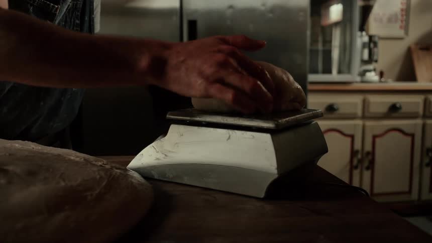 In a dimly lit kitchen a bakers hands carefully place a smooth round portion of dough onto a digital scale to ensure precise measurements before proceeding with the baking process