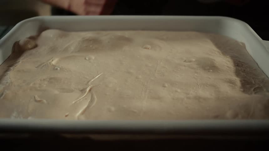 A bakers hands lift the white lid off a large proofing bin revealing a rectangular mass of smooth fermented dough rising with soft air bubbles