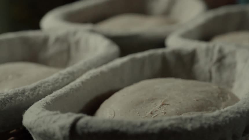 A bakers hands quickly pinch the seam of a rising dough ball resting in a linenlined basket before the entire tray of loaves is lifted and moved away