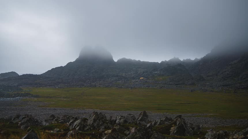 Grass covered field between rocks and mountains under a cloudy sky
