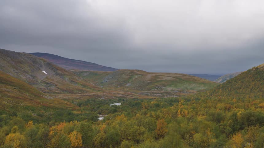 A wide shot captures a vast mountain valley during autumn featuring a winding river flowing through a dense forest of yellowing trees under a heavy overcast sky with rolling hills stretching into the horizon