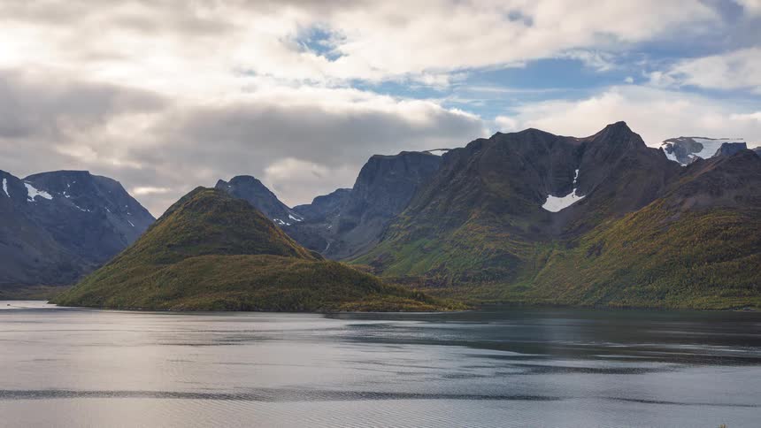 Small boats sail on the water with mountains along the coast under a cloudy sky