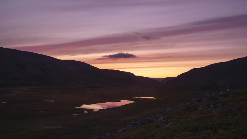 As evening falls over the rugged landscape vibrant streaks of purple and orange clouds drift across the twilight sky mirroring their colors in the still waters of a valley lake