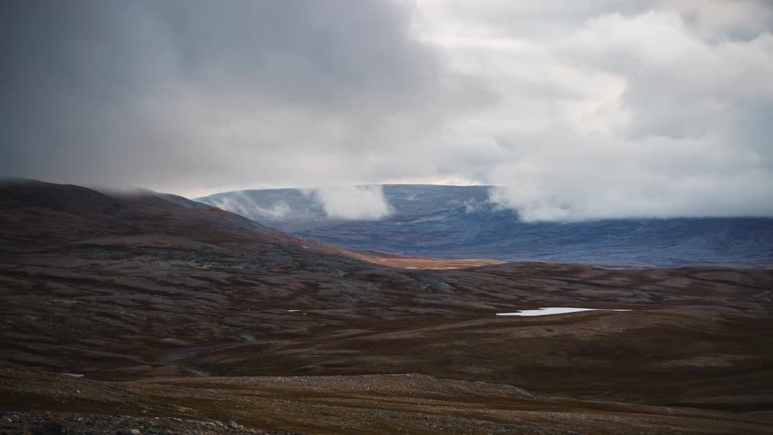 Dark clouds move quickly over a vast desolate mountain range featuring brown rolling hills a small pond in the valley and patches of sunlight hitting the rocky tundra ground