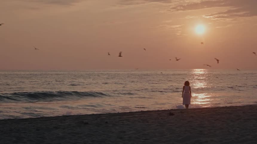 Woman walking on the beach while birds fly through the red sky at sunset stops to stretch